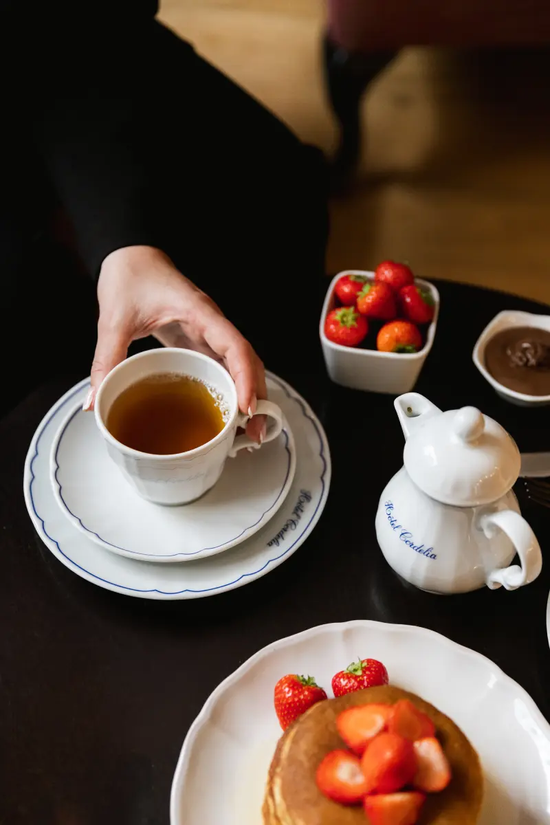 Main saisissant une tasse de the Hotel Cordelia, theiere et pancakes aux fraises sur la table
